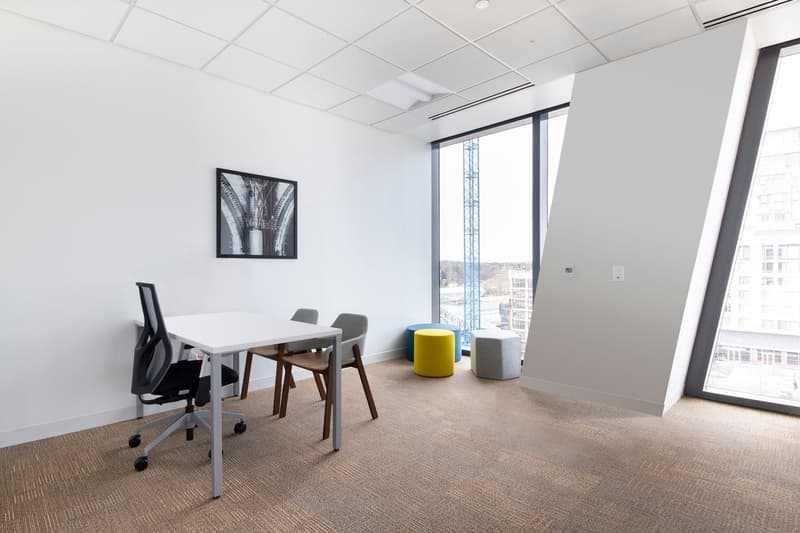 A contemporary meeting room with a white desk, office chairs, blue and yellow stools, and large windows
