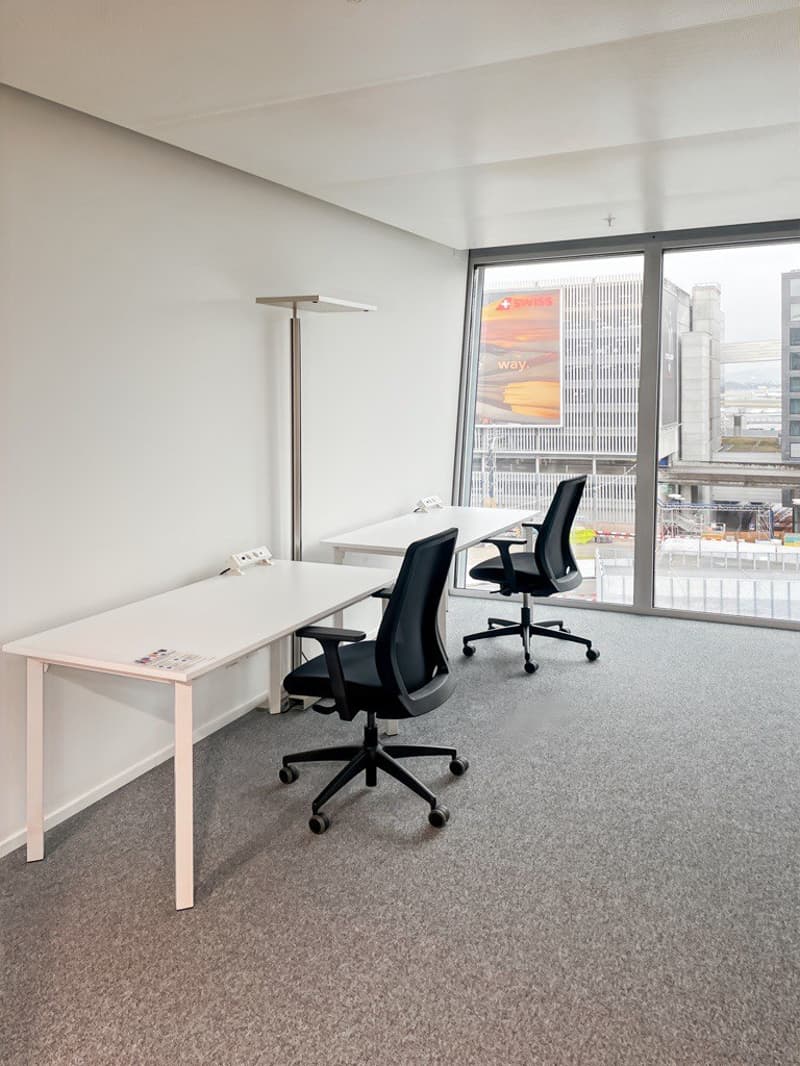 white room with 2 tables and 2 black chairs, grey carpet floor, large windows, lamp