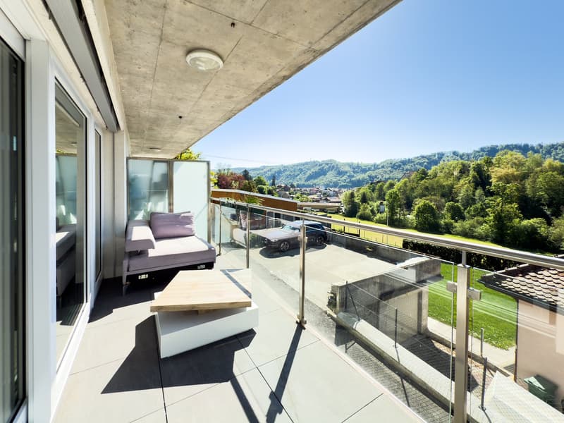 Spacious balcony with glass railing, offering a scenic view of the surrounding hills and greenery. The balcony features comfortable outdoor furniture, including a sofa and a wooden bench, providing a relaxing space to enjoy the natural surroundings.
