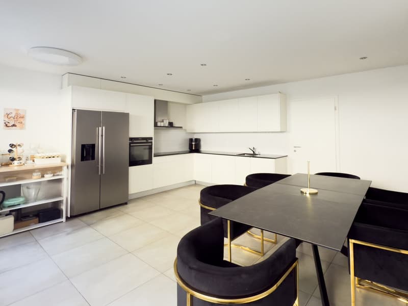 Modern, open-concept kitchen with white cabinets, stainless steel appliances including a refrigerator and oven, a black countertop island with gold-colored stools, and a dining table with black chairs
