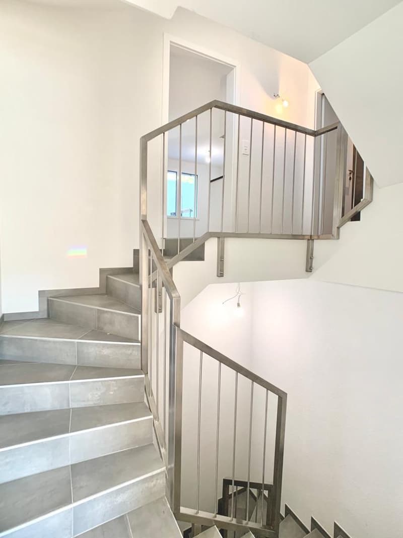 The image shows a modern staircase with a metal railing and steps made of gray tiles. The staircase leads up to a landing with a doorway visible at the top.