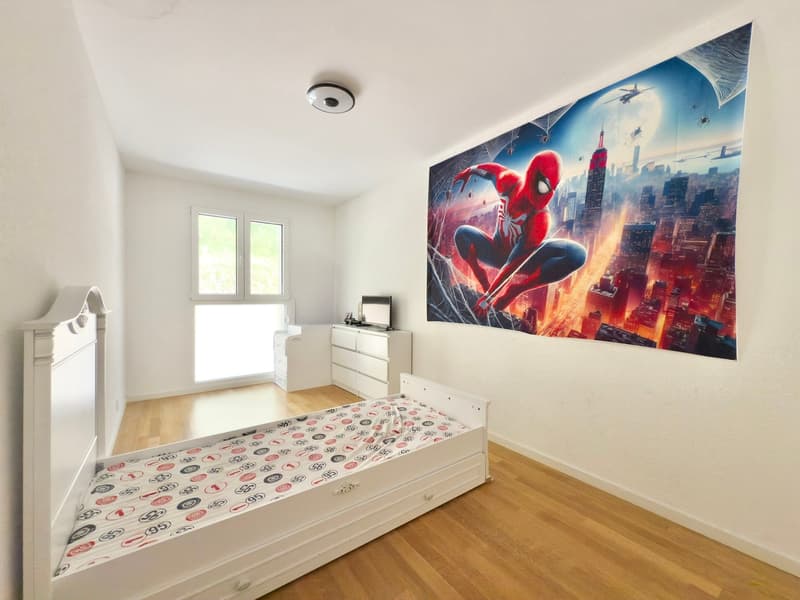 The image shows a bedroom with a large wall mural depicting Spider-Man swinging through a cityscape. The room has white walls, hardwood floors, and a dresser with a TV on top. There is a bed with a patterned bedspread in the center of the room.