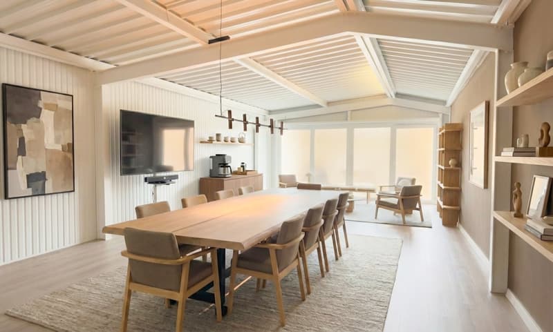 Dining area with long wooden table, beige chairs, modern interior, wooden cabinet, television, coffee maker, large glass windows