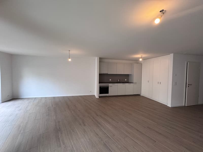 Empty room with wooden floor, white walls, kitchen with sink, cabinets, and oven, and a door.