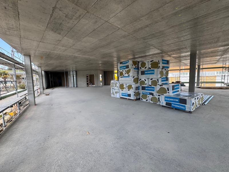 Empty room with concrete floor, high ceiling, construction materials stacked in the corner, metal scaffolding on the left side, and a building under construction in the background.