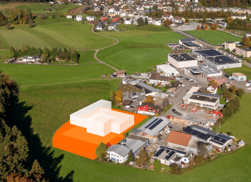 Proposed building outlined on a green field, with many houses and buildings in the background, a few solar panels installed.