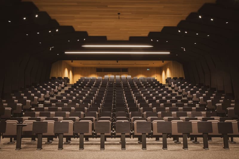Large auditorium with rows of black seats, wooden walls, and overhead lighting fixtures. The stage area is visible at the front of the room.