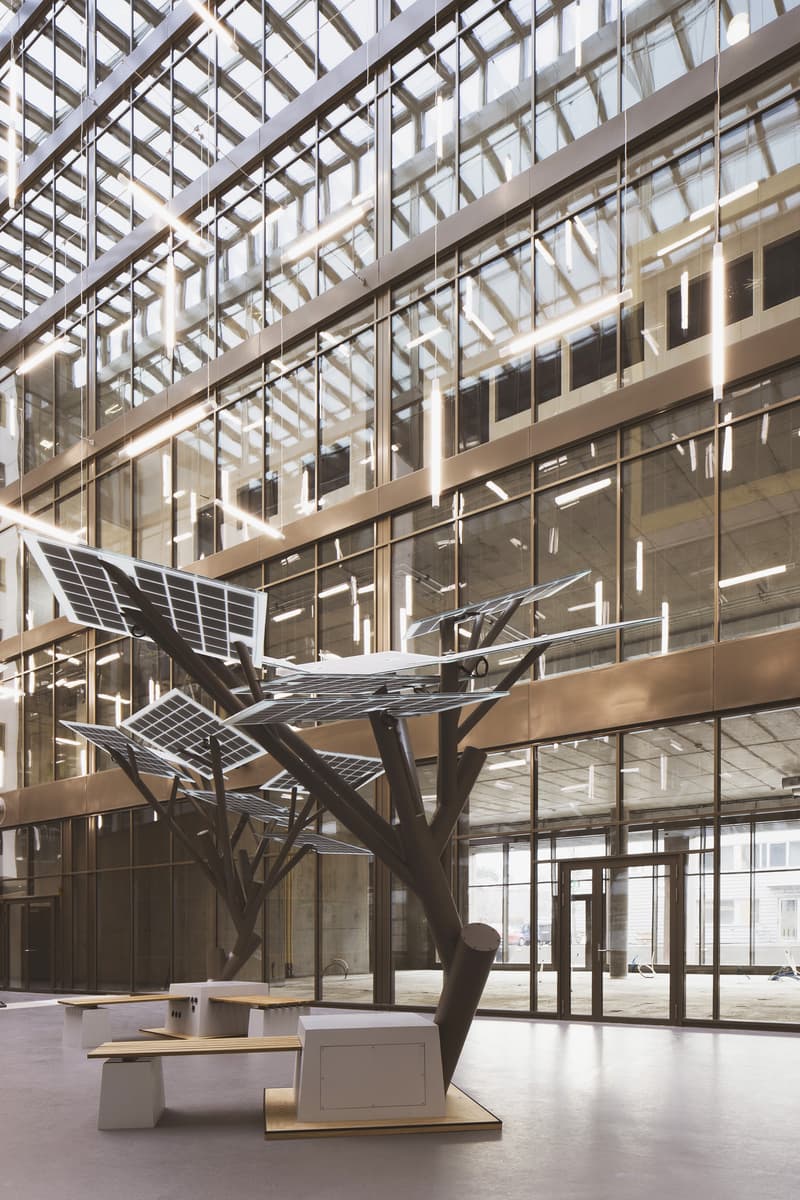 Large open space with glass walls and ceiling, modern architectural design featuring metal and glass structures, sculptural art installation with solar panels in the center of the room