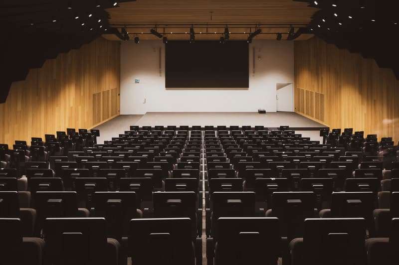 Large auditorium with rows of black theater seats facing a stage with a projection screen. The walls and ceiling are made of wood paneling, and there are spotlights installed overhead.