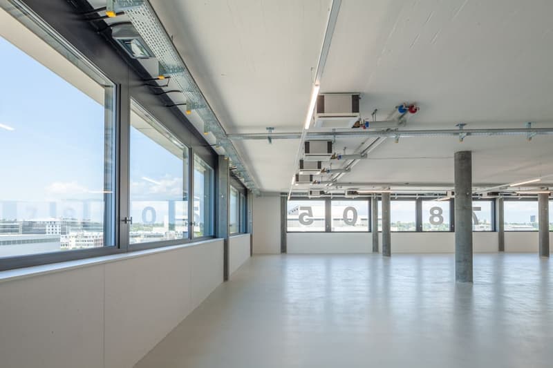 Large open office space with high ceilings, concrete floors, and floor-to-ceiling windows providing natural light. The space has a modern, industrial aesthetic with exposed building systems and signage on the walls.