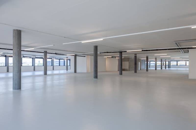 Large open floor plan with concrete columns, exposed ceiling, and large windows providing natural light. The space appears to be a commercial or office area ready for customization and use.