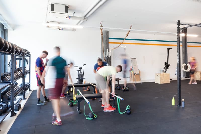 The image shows a gym or fitness center with various exercise equipment and people working out. The space has white walls with colorful stripes, and the floor is covered in black mats. There are weight racks, resistance bands, and other fitness tools visi