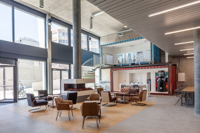 Large open-concept office space with concrete walls and ceiling, large windows, leather armchairs and sofas, and a spiral staircase leading to a mezzanine level.