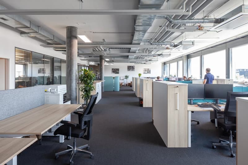 Open-plan office space with cubicles, desks, computers, and a dishwasher visible. The ceiling has exposed ductwork and the walls have glass partitions. There are plants and other decor elements throughout the space.