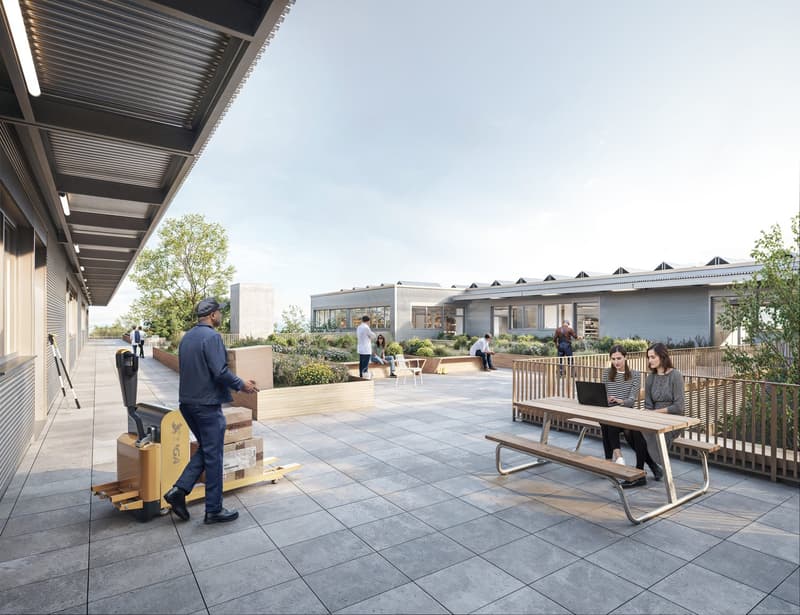 Outdoor patio area with benches, tables, and landscaping. People are seated and working on laptops, suggesting it is a communal workspace or gathering area.