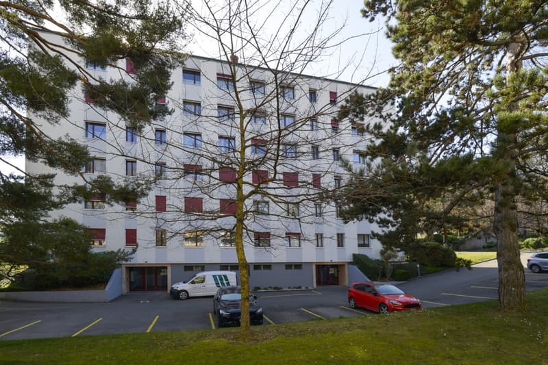 Apartment building, several floors, windows with red shutters, cars parked, paved parking lot, trees