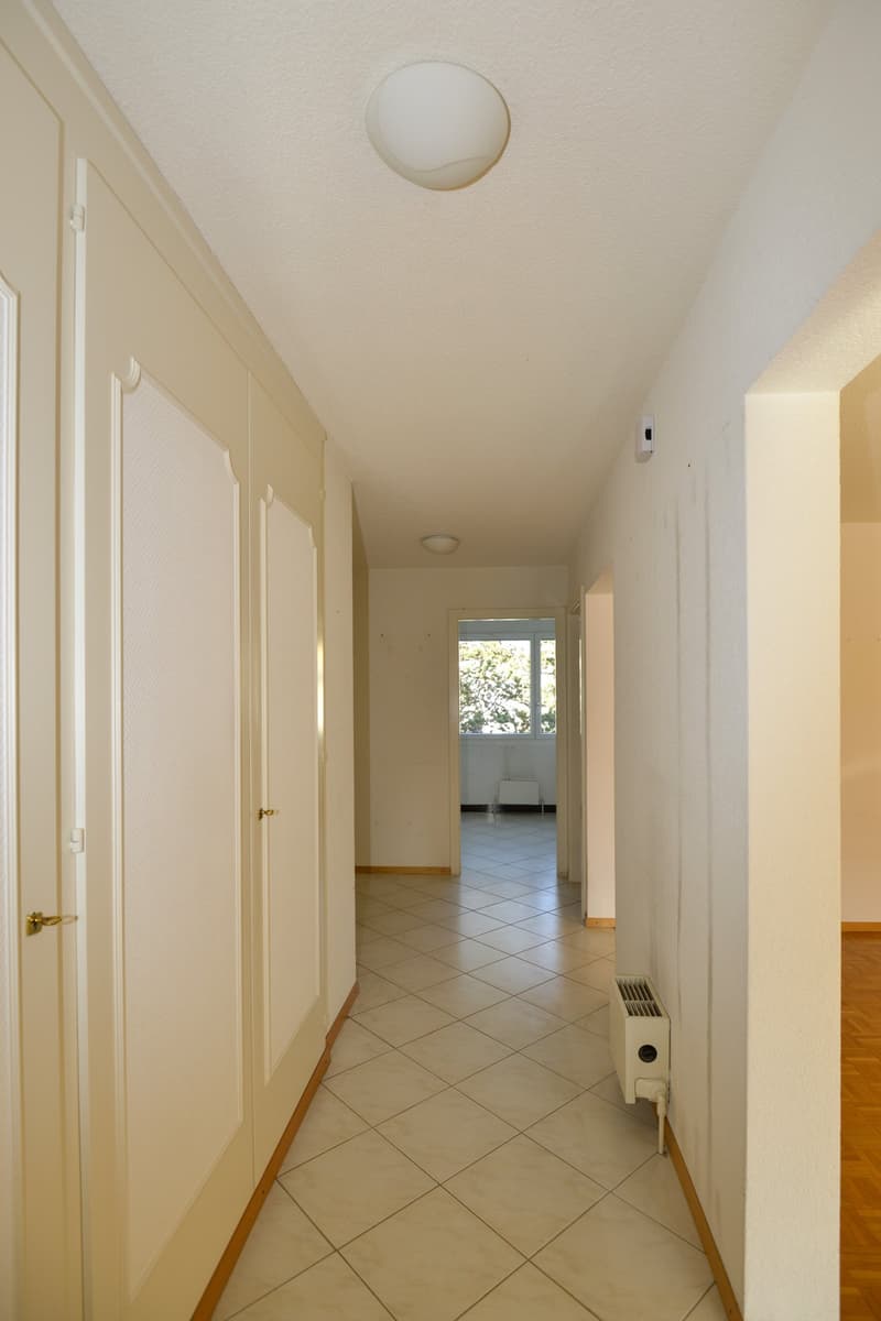 White walls, tile flooring, two white doors, ceiling lamps, small window with greenery outside