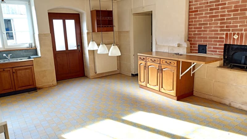 Spacious kitchen with wooden cabinets, tile flooring, brick walls, and a fireplace. The room has large windows and pendant lighting fixtures.
