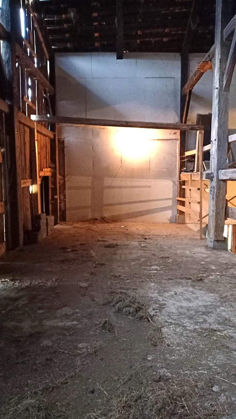 The image shows the interior of an old, wooden barn. The space is dimly lit, with a wooden frame structure and a dirt floor. There are various wooden pallets and other storage items visible in the background.