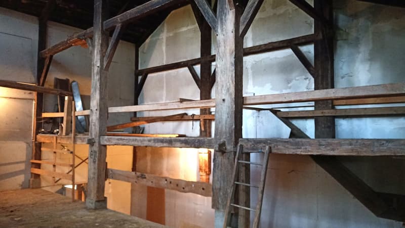 The image shows an old, rustic attic space with exposed wooden beams and shelves. The space appears to be used for storage, with various items and boxes visible on the shelves. The overall atmosphere is dark and dimly lit, giving the space a vintage and a