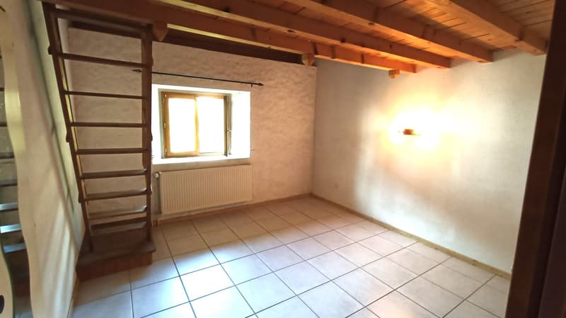This image shows a room with a wooden ceiling and beams, a window, and tiled flooring. The room appears to be empty, with a ladder-like structure against one wall.