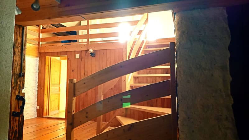 The image shows the interior of a wooden cabin or chalet. The walls and ceiling are made of wooden panels, and there is a wooden staircase leading to an upper level. The room appears to be a living or common area, with a yellow door visible in the backgro