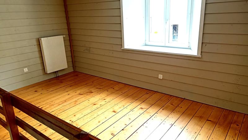 This image shows a room with wooden walls and a hardwood floor. There is a radiator on the wall and a large window that provides natural light. The room appears to be empty, with no furniture or other objects visible.
