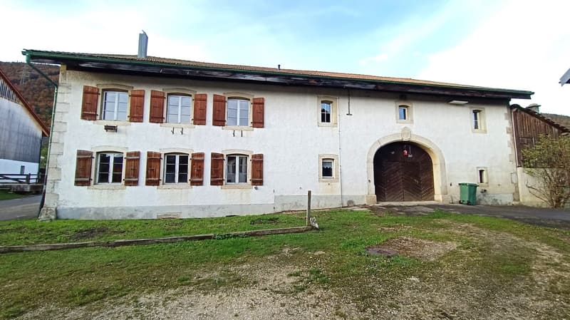 Two-story building with white walls, red tile roof, and wooden shutters. The building has a large arched entrance and a garage door on the ground floor.