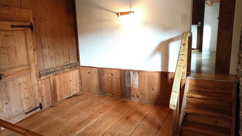 This image shows a rustic interior space with wooden walls and floors. The room has a wooden door and shelving units along the walls. The lighting in the room appears to be provided by a wall-mounted light fixture. The overall aesthetic is that of a tradi