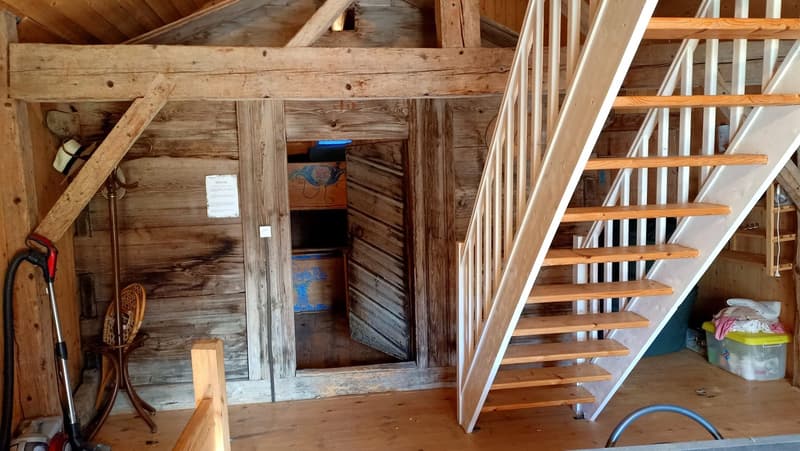 The image shows the interior of an attic space. The walls are made of weathered, wooden boards, and there is a wooden staircase leading up to the attic. The attic appears to be used for storage, with various items and boxes visible.