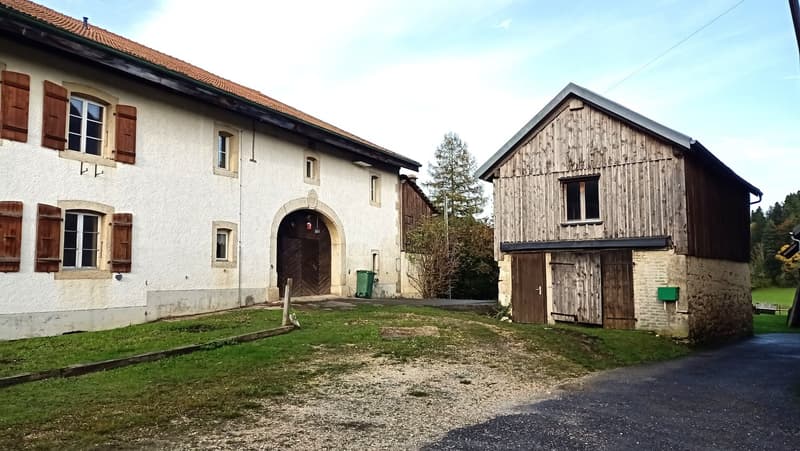 Two-story farmhouse with white walls and wooden shutters, a wooden barn structure, and a grassy yard with a gravel driveway
