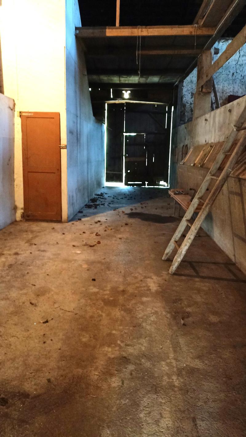 The image shows a dimly lit, cluttered attic or storage space. There is a wooden door visible, along with various wooden beams and debris on the floor. The space appears to be unfinished and used for storage or renovation purposes.