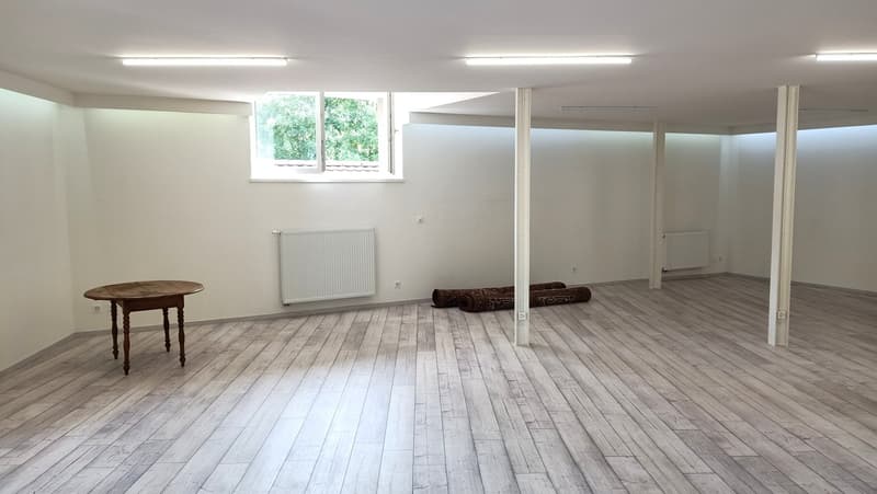 white walls, ceiling with multiple lights, white wood floor, small table, two rolled mats, wall-mounted radiators, window
