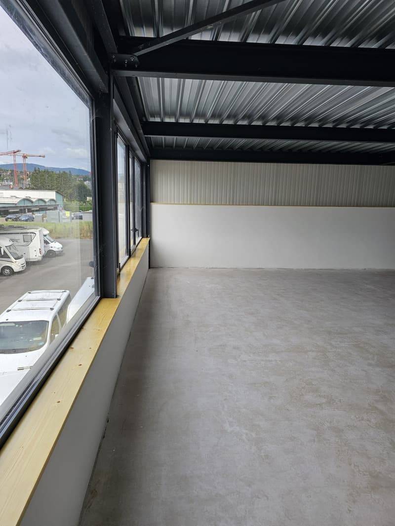 The image shows the interior of a warehouse or industrial space. The space has a high ceiling with black metal beams and a corrugated metal roof. The walls are lined with white panels, and the floor is a smooth concrete surface. There are large windows al
