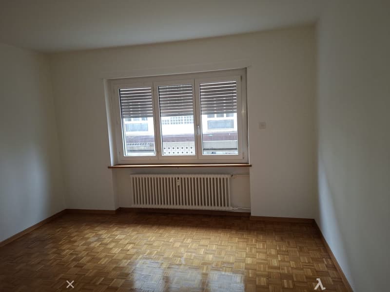 Empty room with large window, radiator, wooden floor, white walls.