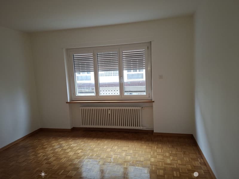 Empty room with large window, radiator, wooden floor, white walls.