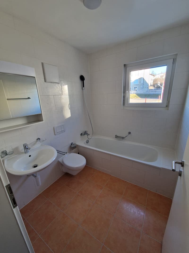 Bathroom with white tiles, white sink, white bathtub, white toilet, mirror, window, and brown tiles on the floor.