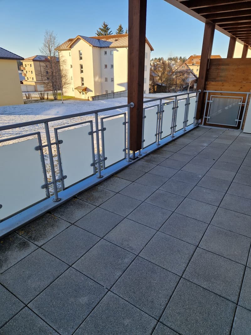 Balcony with metal railings and tiled floor, overlooking a snowy landscape with buildings in the background.