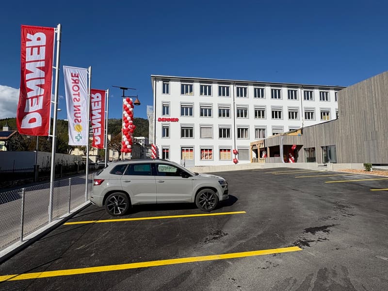 A multi-story commercial building with a large parking lot in front. The building has white exterior walls and several windows. There are red and white flags or banners displayed outside the building, indicating it is a commercial or retail establishment.