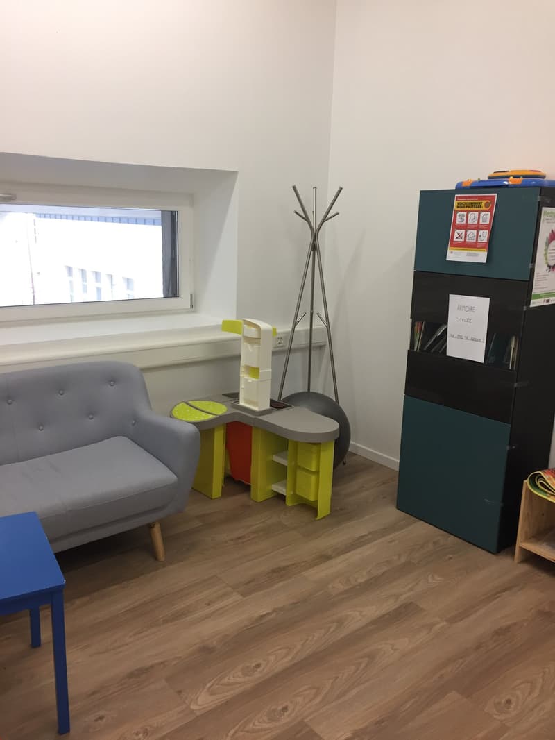 The image shows a room with a gray sofa, a small yellow table, and a black storage unit. There is a window and a coat rack with a hanging umbrella. The room has a wooden floor and white walls.