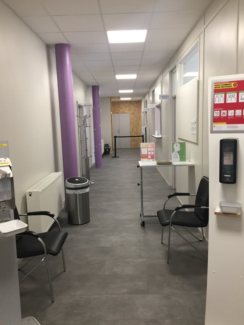 The image shows a hallway or office space with gray tiled flooring, white walls, and recessed lighting. There are several chairs and a table with a dishwasher visible. The space appears to be a professional or commercial setting, possibly a medical or adm
