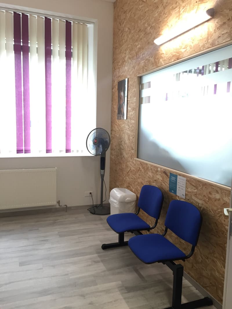 The image shows an office space with a whiteboard, blue office chairs, and vertical blinds covering a window. The walls are made of OSB (oriented strand board) panels, and there is a fan visible in the room.