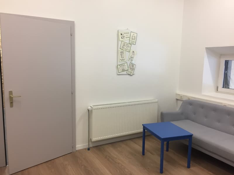 The image shows a bedroom with a gray tufted sofa, a blue side table, a radiator, and a white door. The walls are white, and the floor is made of hardwood. There is a bulletin board on the wall with various notes and drawings.