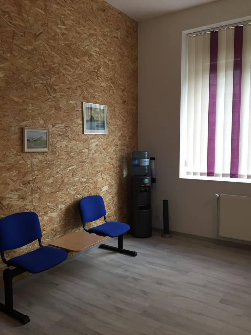 The image shows a small office space with an OSB (oriented strand board) wall, two blue chairs, a water dispenser, and framed artwork on the wall. The room has a vertical window with purple curtains and a laminate wood floor.