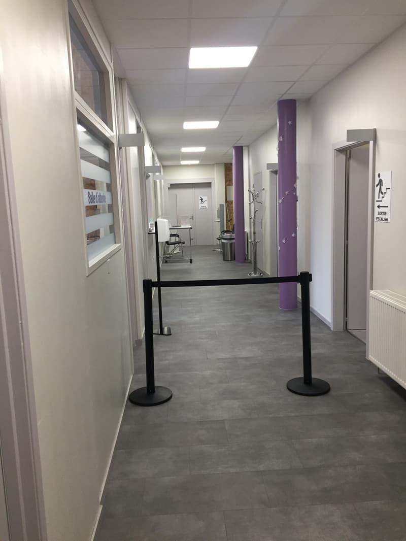 The image shows a hallway or corridor in an office or commercial space. The walls are painted in a light color, and the floor is covered with gray tiles. There are several doors and rooms visible along the hallway, and the lighting is provided by recessed