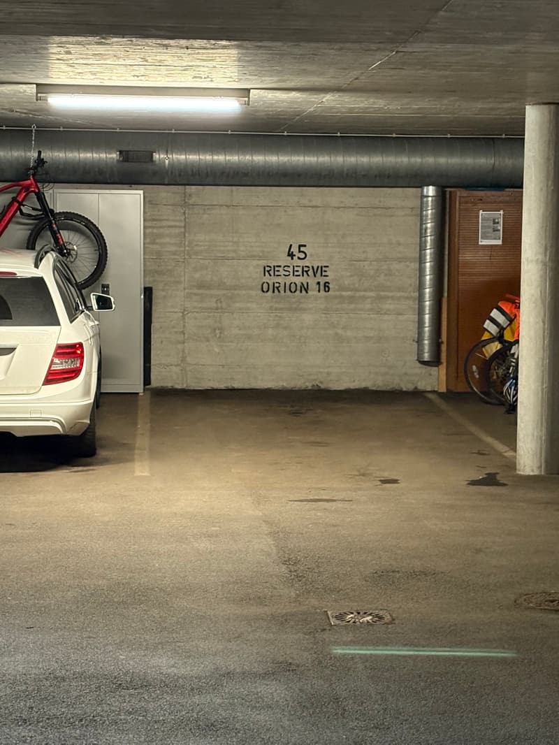 Concrete walls and floor, reserved parking spot labeled 'RESERVE ORION 16', white car and motorcycle parked in the garage