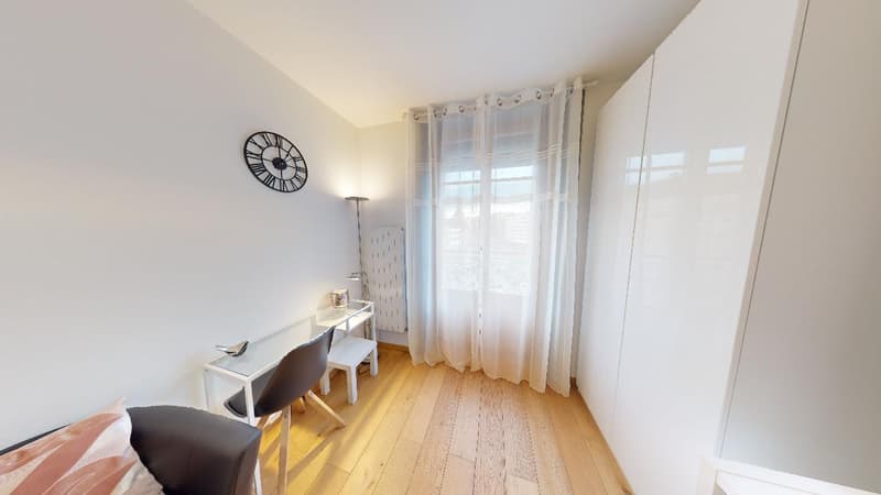 Spacious bedroom with large windows, hardwood flooring, white walls, a desk with chairs, and a decorative wall clock.