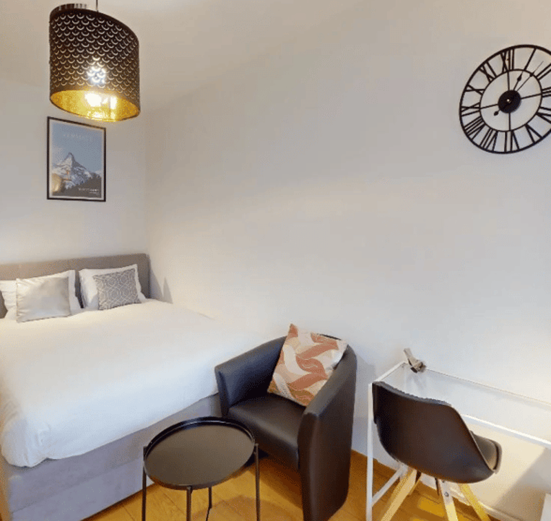 Bedroom with bed, chair, and desk. White walls, wooden floor, framed picture, and round clock on the wall.