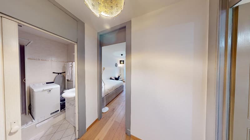 The image shows the interior of an apartment. It has a hallway with a washing machine visible in a bathroom, and a bedroom visible through an open doorway. The apartment has hardwood floors and white walls, with a chandelier in the hallway.