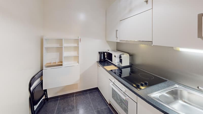 Compact kitchen with white cabinets, black countertop, built-in appliances including a microwave and coffee maker, and a small storage shelf on the wall.